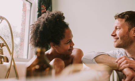 A woman takes a hot bath as her partner rests his arm on the side of the tub and gazes at her lovingly.